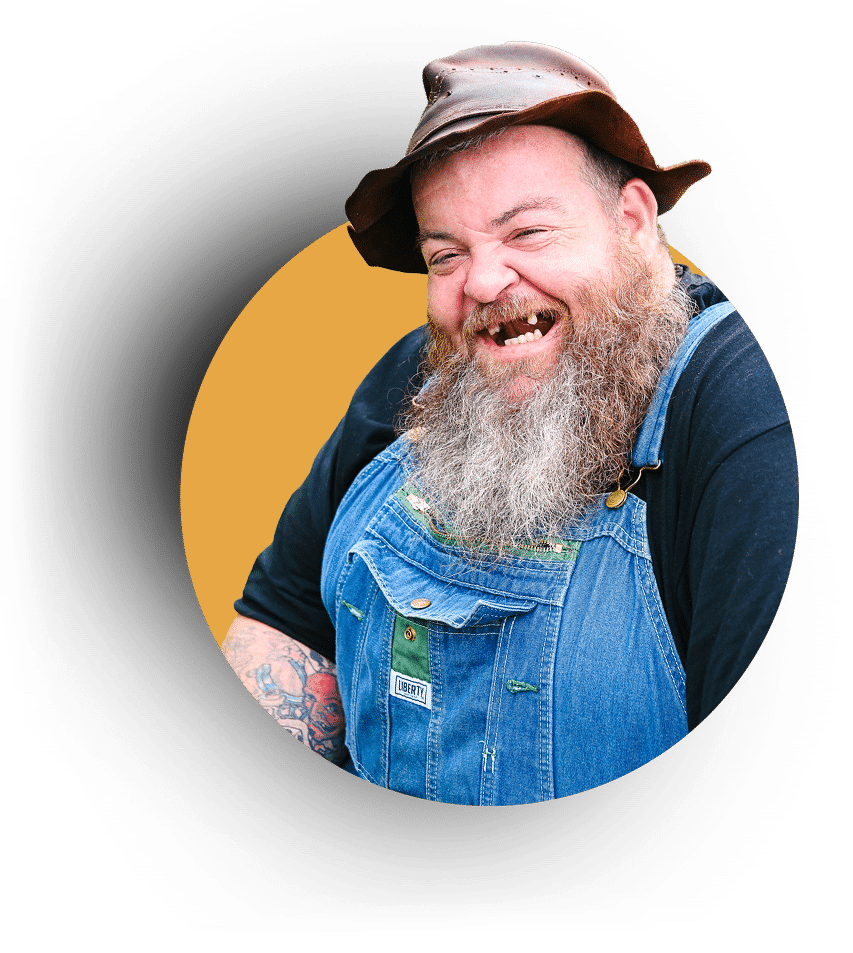 Bearded man wearing a brown hat, denim overalls, and a black shirt, smiling broadly against an orange background.