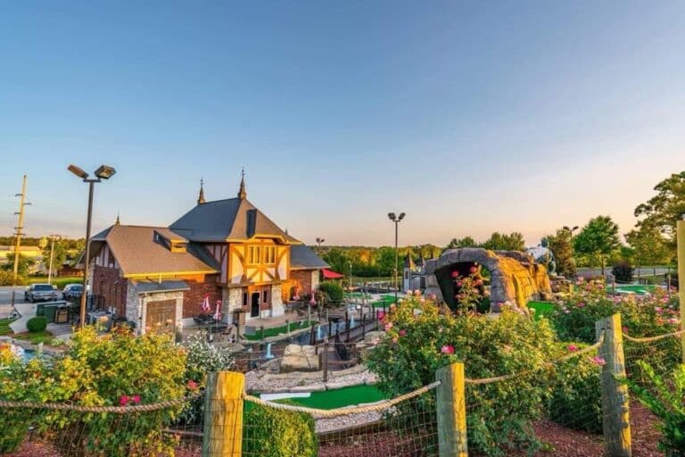 A mini-golf course with landscaped greenery, a castle-themed building, and rock formations under a clear sky at sunset.