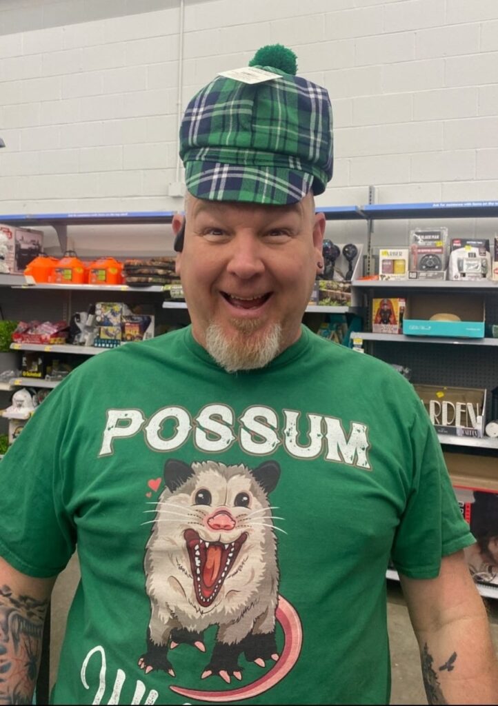 A man in a green "POSSUM" T-shirt and a green plaid hat stands in a store aisle, smiling at the camera. Shelves with various products are visible in the background.