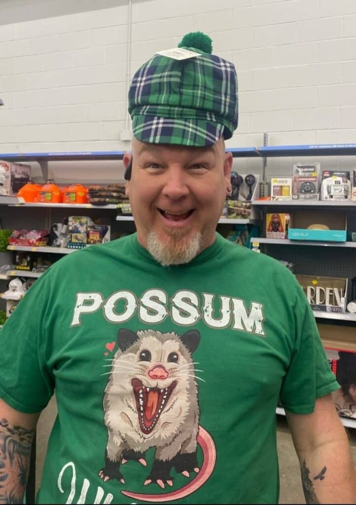 A man standing in a store aisle wears a green plaid hat and a green shirt with a cartoon possum and the word "POSSUM" on it. He is smiling at the camera.