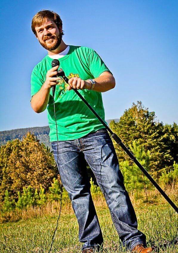 A man with a beard stands outside on grass, holding a microphone and stand, wearing a green t-shirt and jeans, with trees and blue sky in the background.