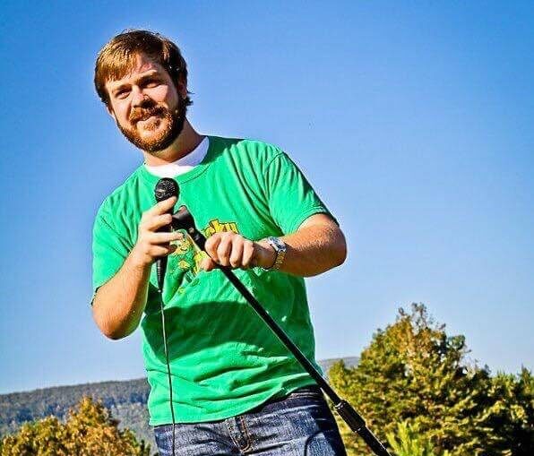 A man in a green T-shirt and jeans stands outdoors on grass, holding a microphone and stand, with trees and blue sky in the background.