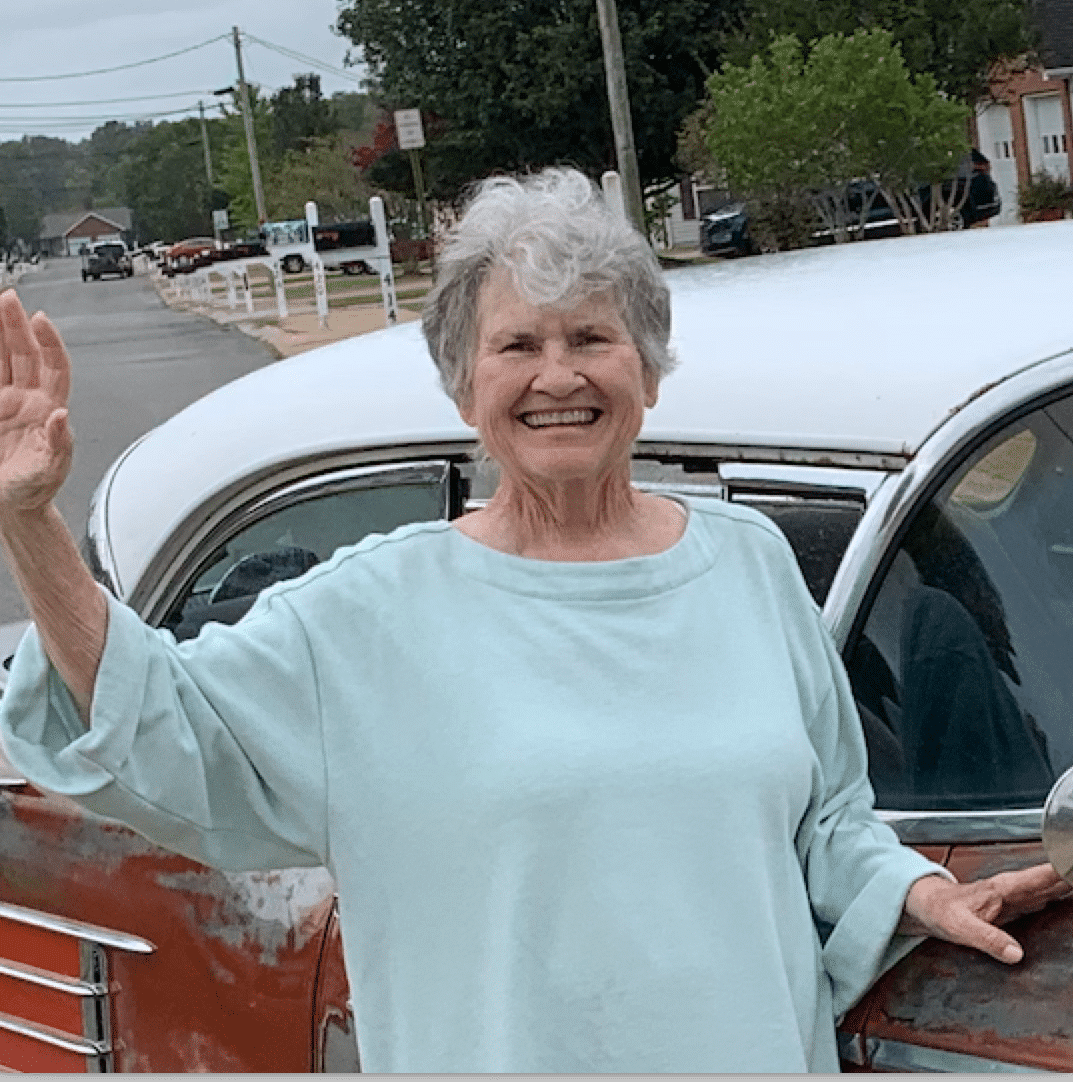 An older woman with short gray hair smiles and waves while standing next to a vintage car parked on a street.